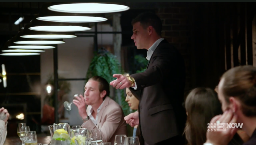 michael stands up pointing during a dinner party as the others remain seated, mafsau season 7
